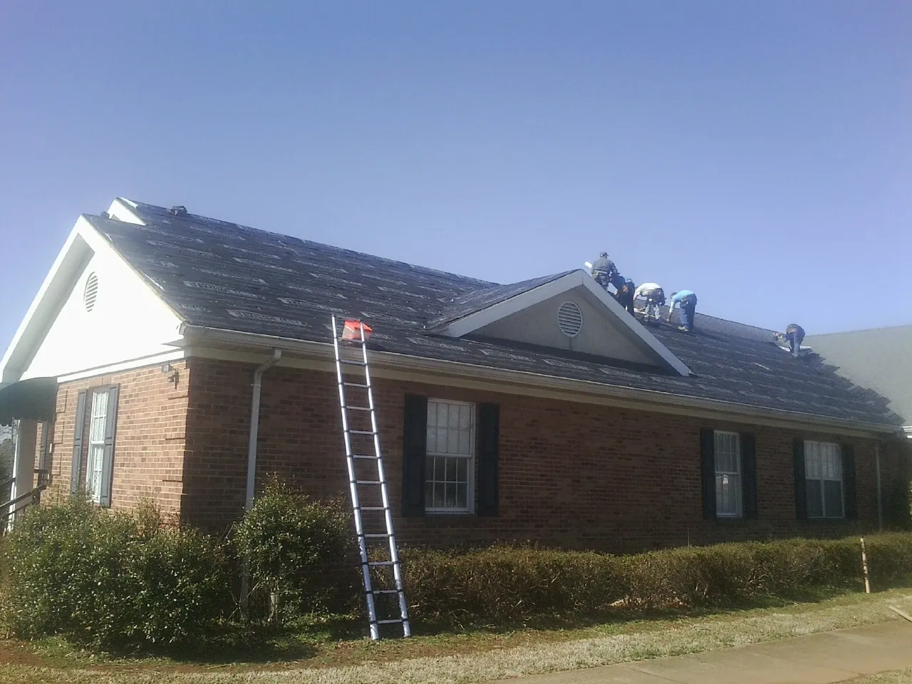 Crew tearing shingles off a brick ranch roof with a ladder set — Emergency Roof Repair work by our crew in Pennellville