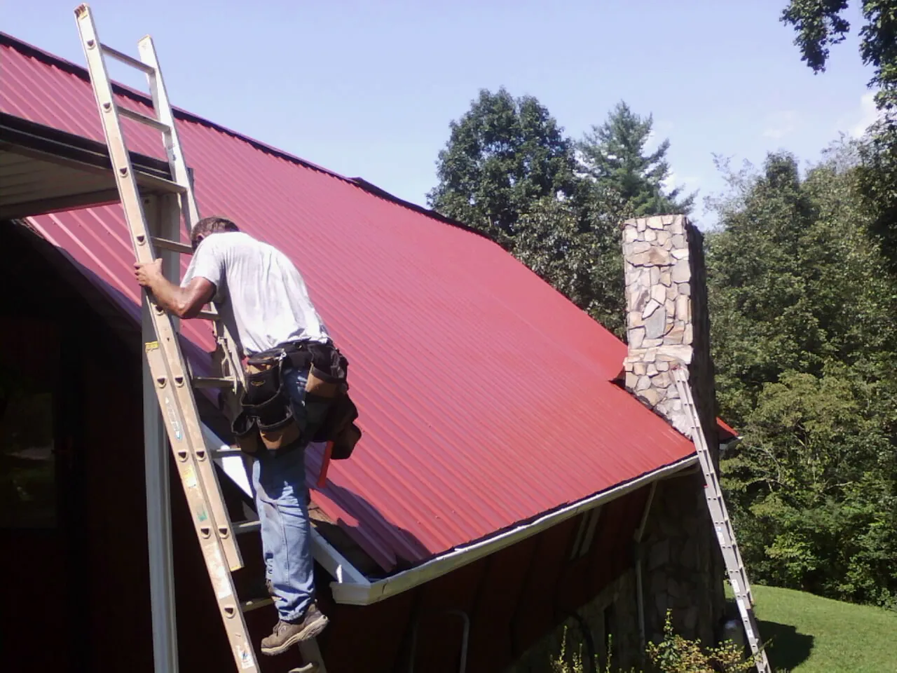 Roofer on a ladder beside a red metal roof and stone chimney — Soffit & Fascia Repair by Roofing Brothers in Pennellville